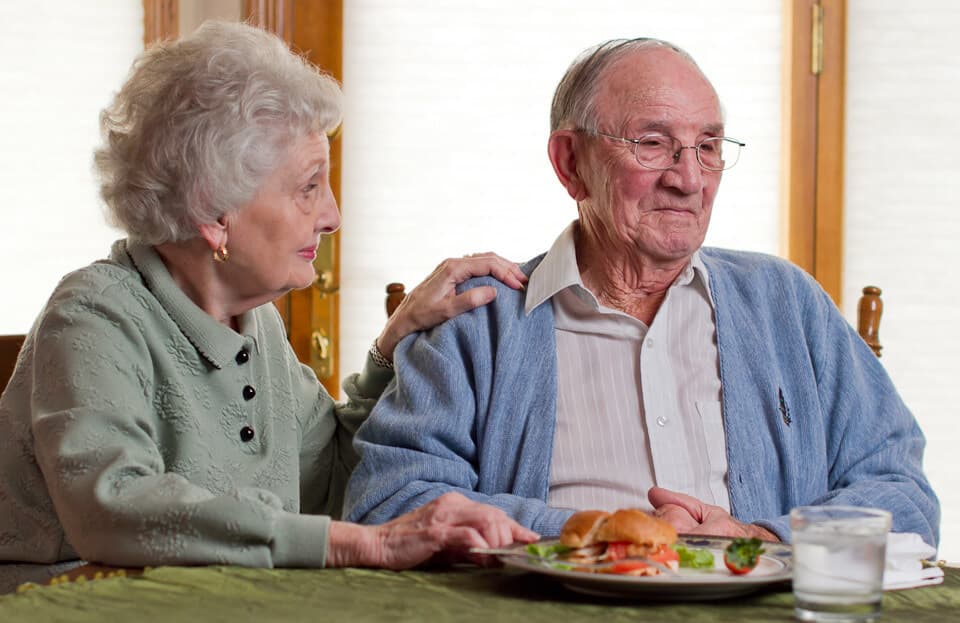 An elderly woman comforts an elderly man at a dining table with a sandwich, salad, and a glass of water. - Home Instead