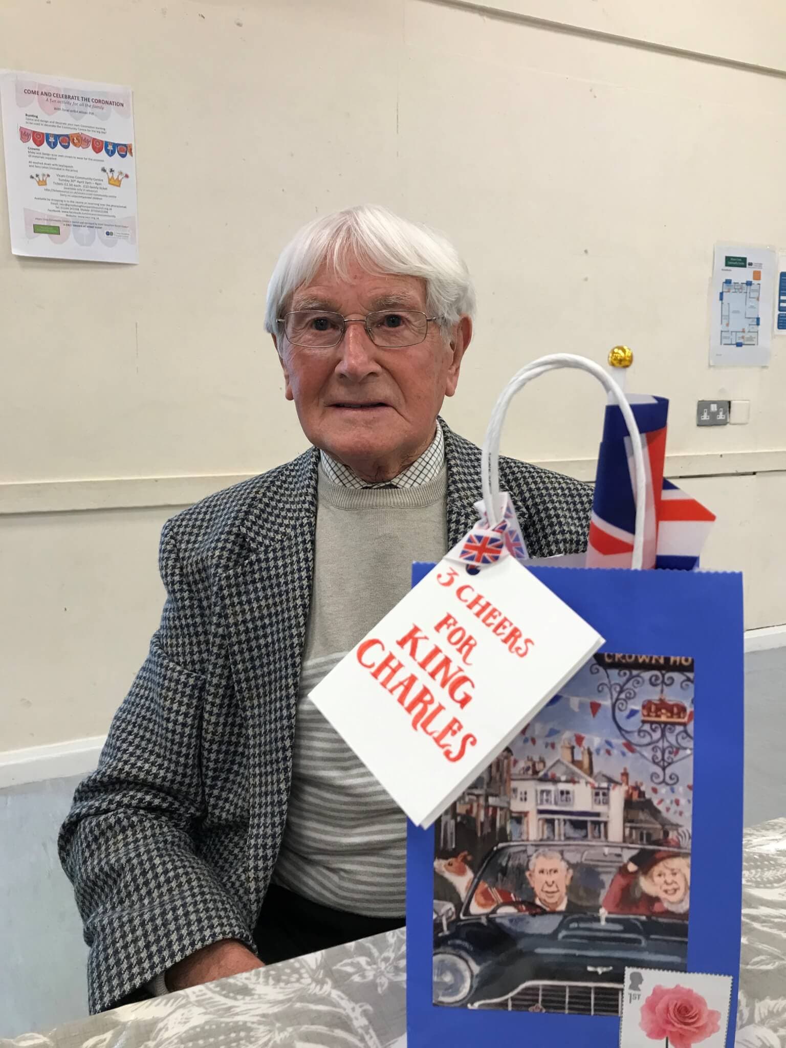 An elderly man sitting at a table with a gift bag labeled "3 CHEERS FOR KING CHARLES" and a small British flag. - Home Instead