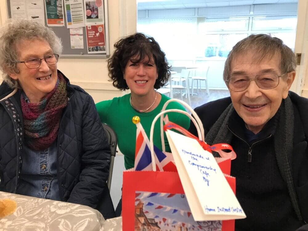 Three adults smiling indoors, a gift bag with small British flags and a tag in front of them. Bulletin board in background. - Home Instead