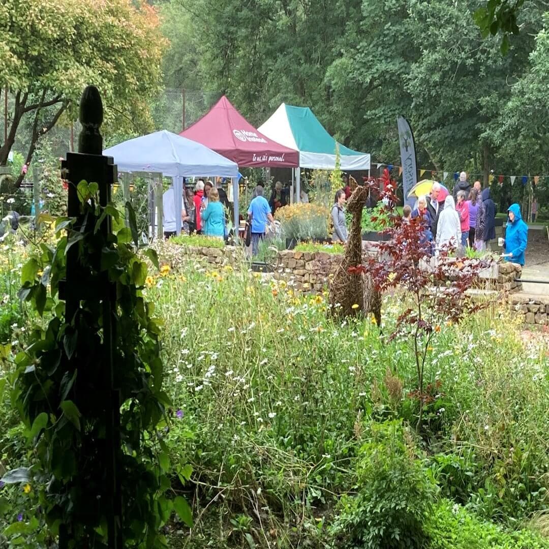 People gathered at an outdoor event with colorful tents and lush greenery in a garden setting. - Home Instead