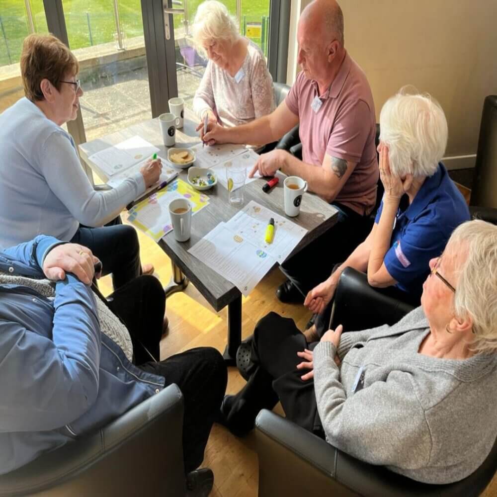 Six elderly people sitting around a table, engaged in discussion with papers and cups of coffee in front of them. - Home Instead