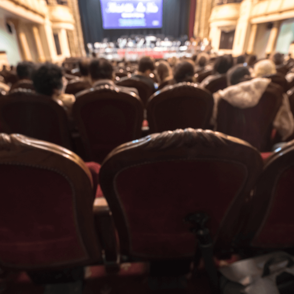 Blurred view from the back of an audience seated in a theater, focusing on a stage with a large screen above it. - Home Instead