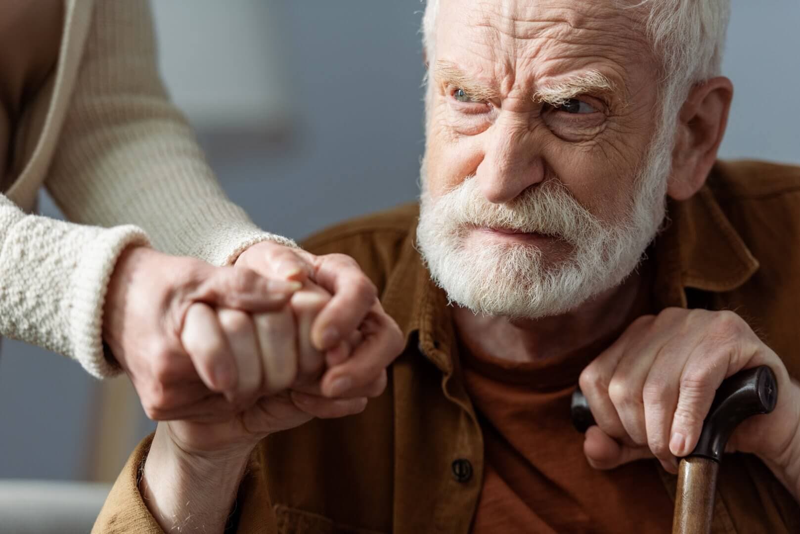 Elderly man with a cane grips someone's hand, looking intently with a serious expression. - Home Instead