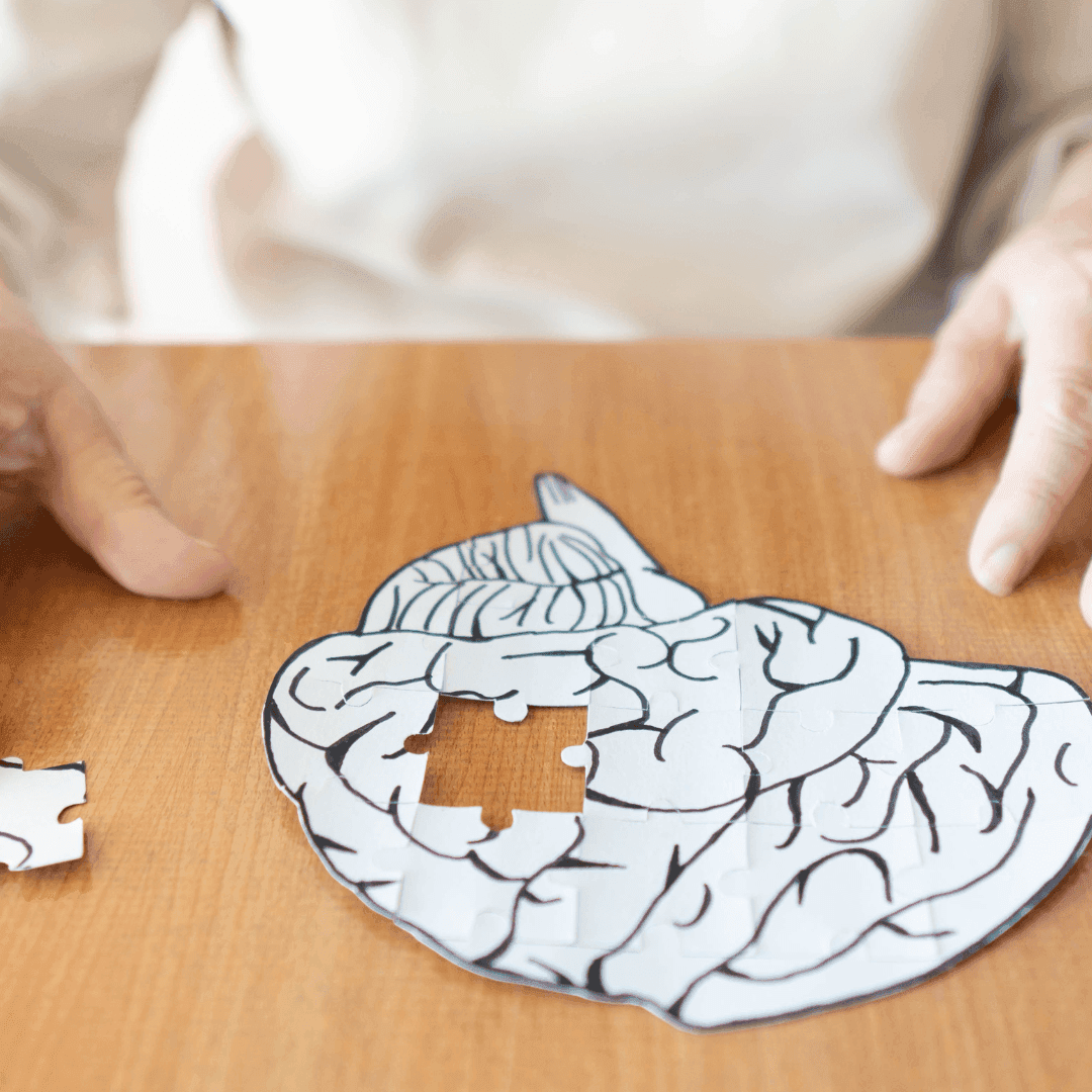 A person working on a brain-shaped puzzle with a missing piece on a wooden table. - Home Instead