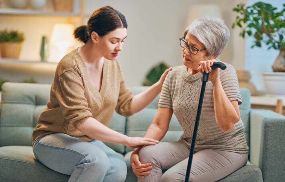 Young woman comforting an elderly woman with a cane, sitting together in a cozy living room. - Home Instead Bournemouth & Christchurch