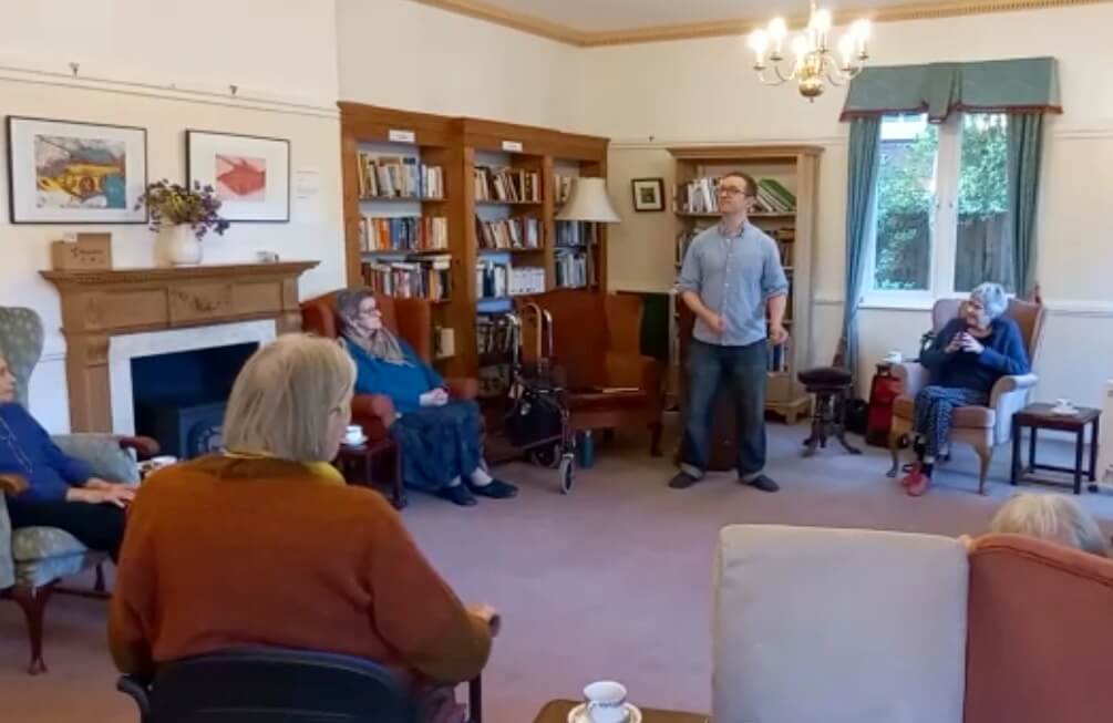 A small group of elderly individuals are gathered in a room while a man stands and speaks to them. - Home Instead