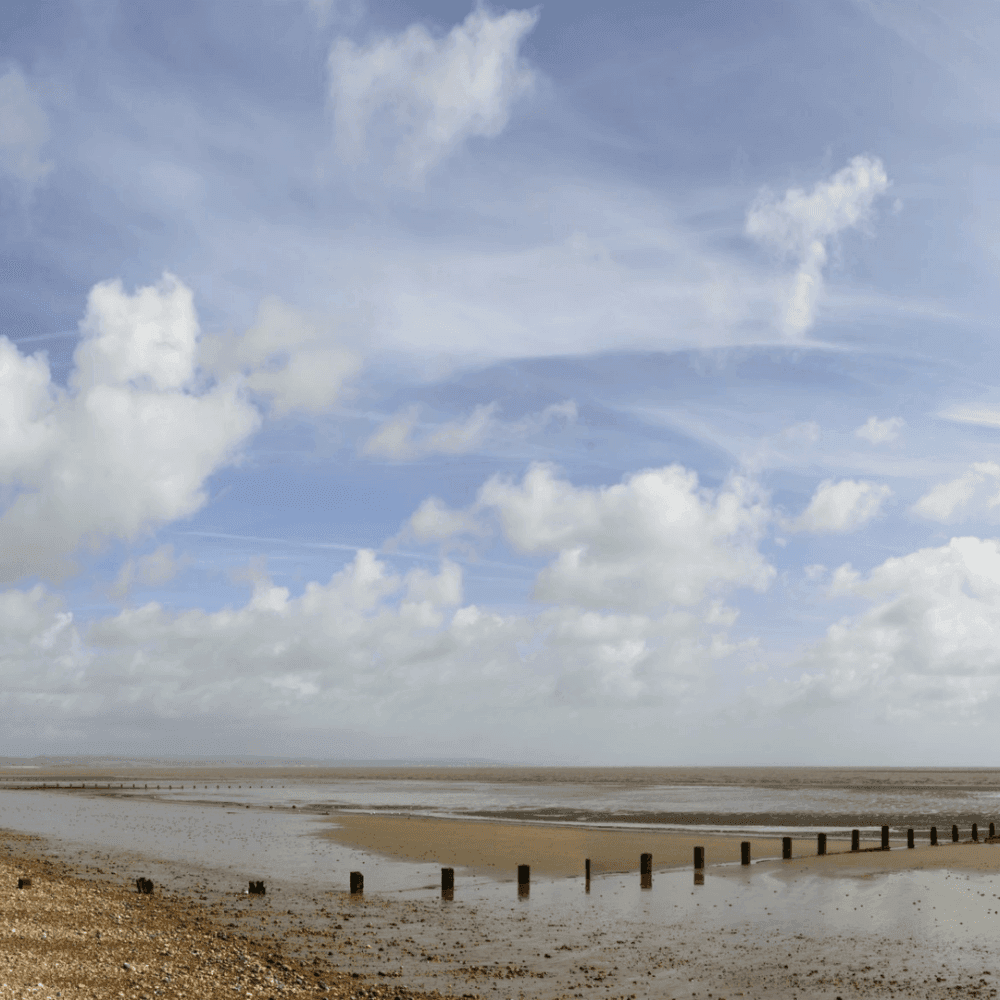 A sandy beach under a cloudy sky with wooden posts leading out into the water. - Home Instead