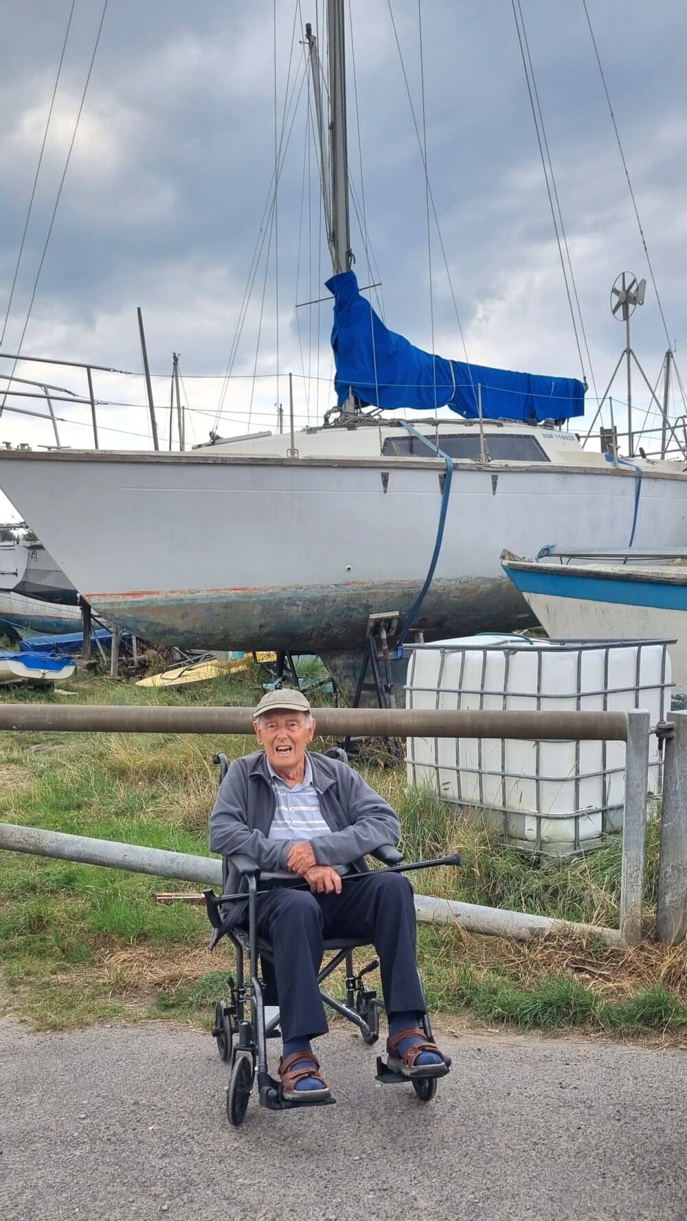 An elderly man sits in a wheelchair, smiling near a large sailboat with a blue cover, under a cloudy sky. - Home Instead