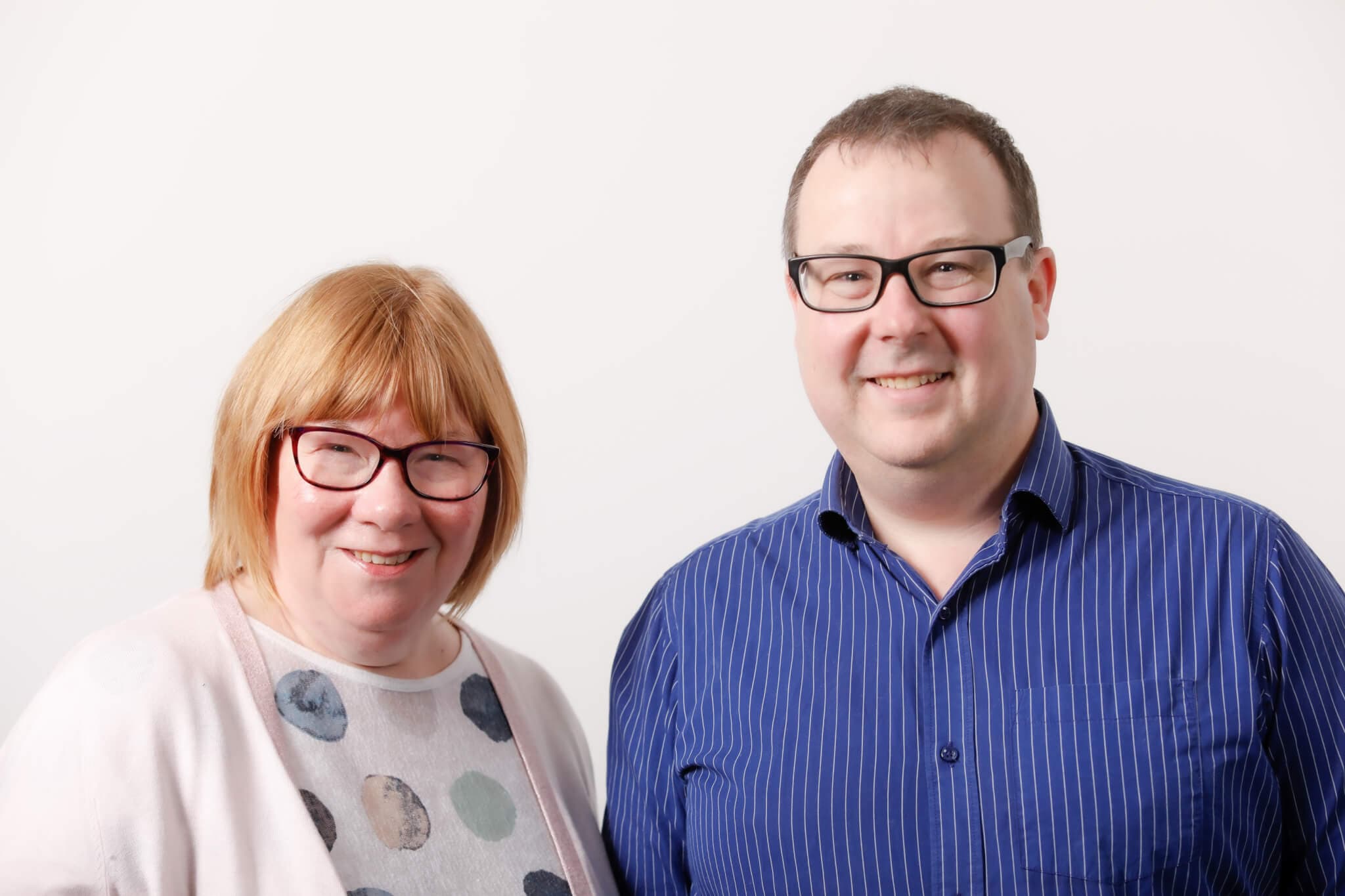 Two people, both wearing glasses, smile in front of a plain white background. One is in a polka dot shirt, the other in blue stripes. - Home Instead