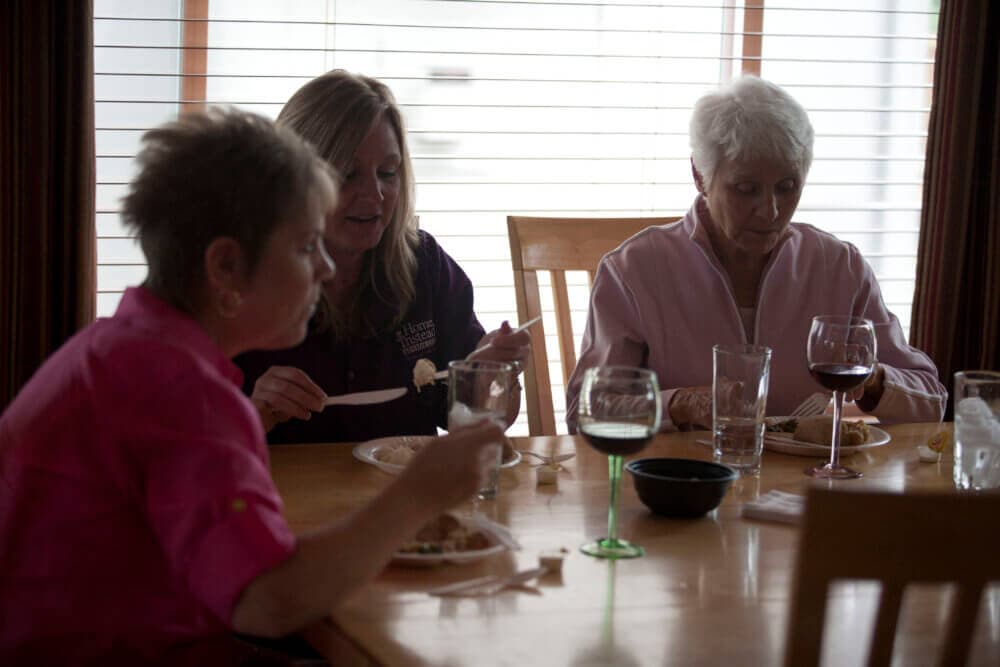 Three women eating and drinking wine at a dining table in front of a window with blinds. - Home Instead