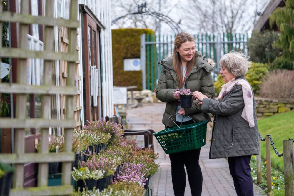 Two women holding flower pots and shopping together in an outdoor plant nursery. One has a shopping basket. - Home Instead