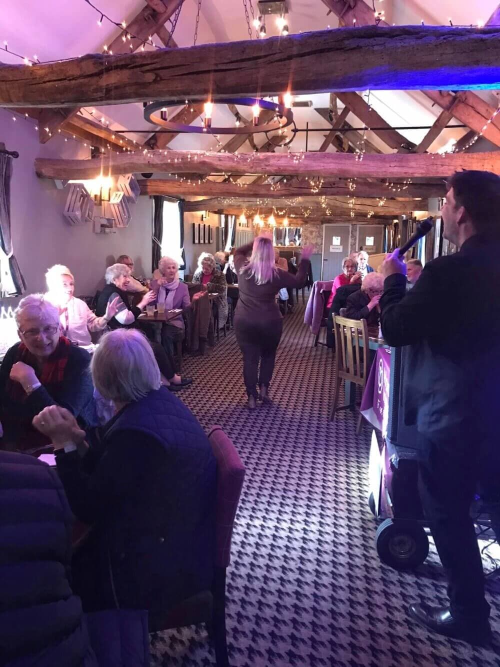 A lively, indoor gathering with people seated at tables, a singer performing, and a woman dancing in the aisle. - Home Instead