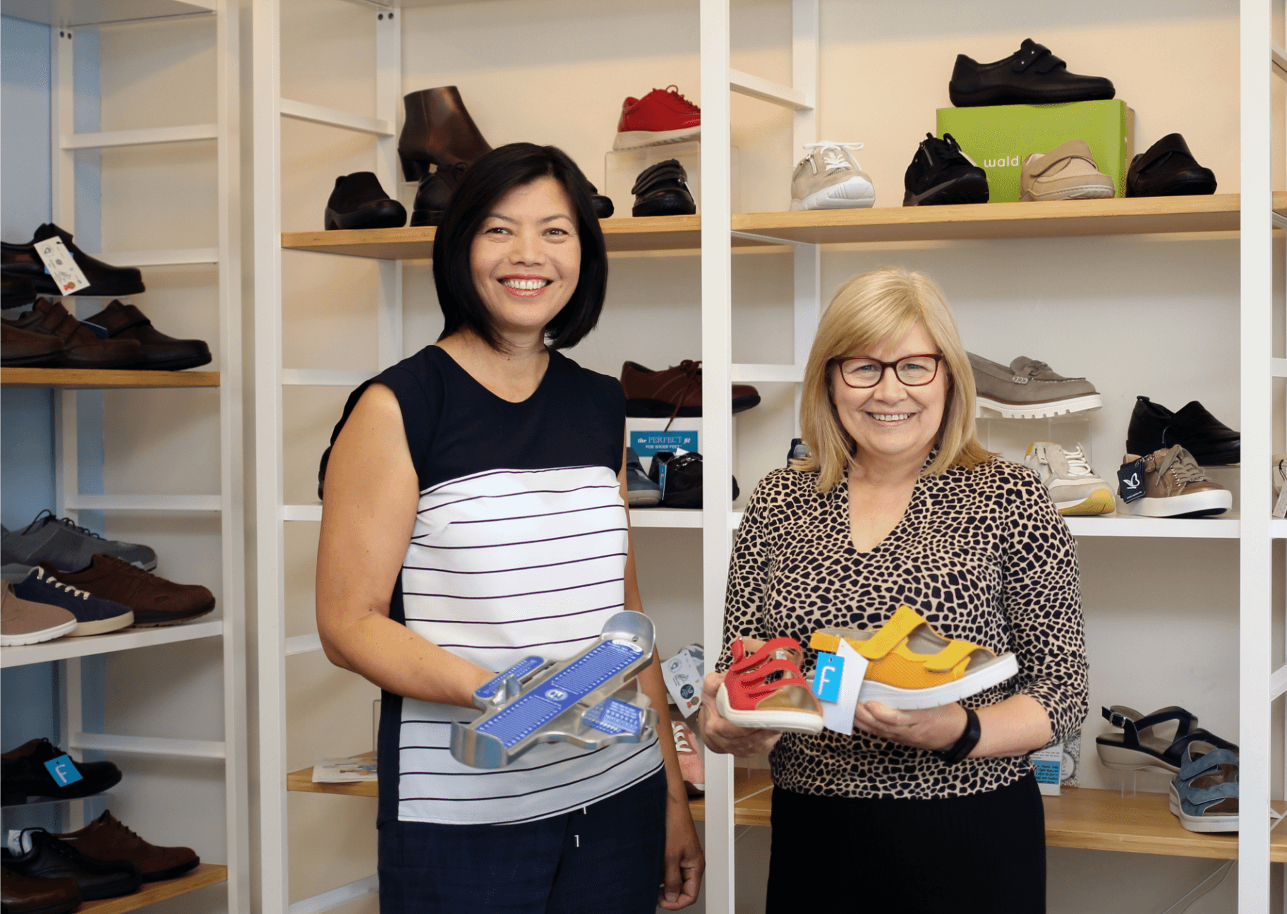 Two women holding shoes, standing in a shoe store with various shoes displayed on shelves behind them. - Home Instead