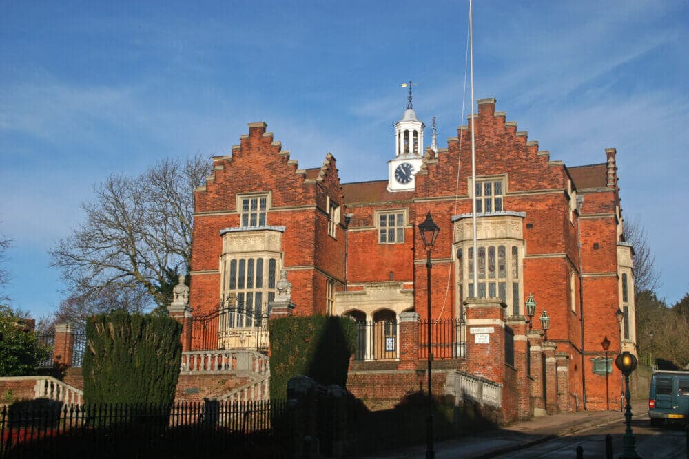 Historic red brick building with a clock tower, gabled roofs, and lush green hedges lining the path on a sunny day. - Home Instead