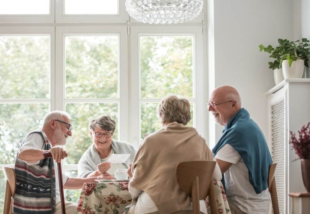 Four smiling elderly people seated around a table, engaged in conversation in a sunlit room with large windows. - Home Instead Southampton