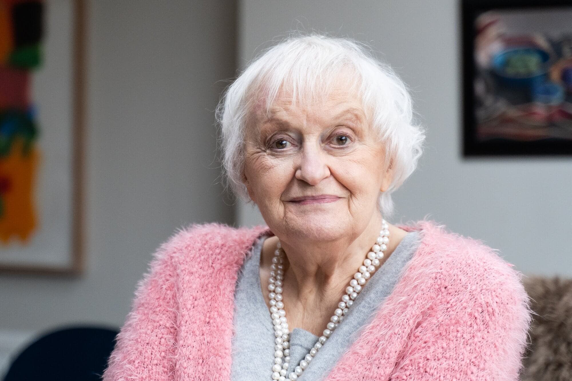 Elderly woman with white hair wearing a pink sweater and pearl necklace, smiling gently indoors. - Home Instead