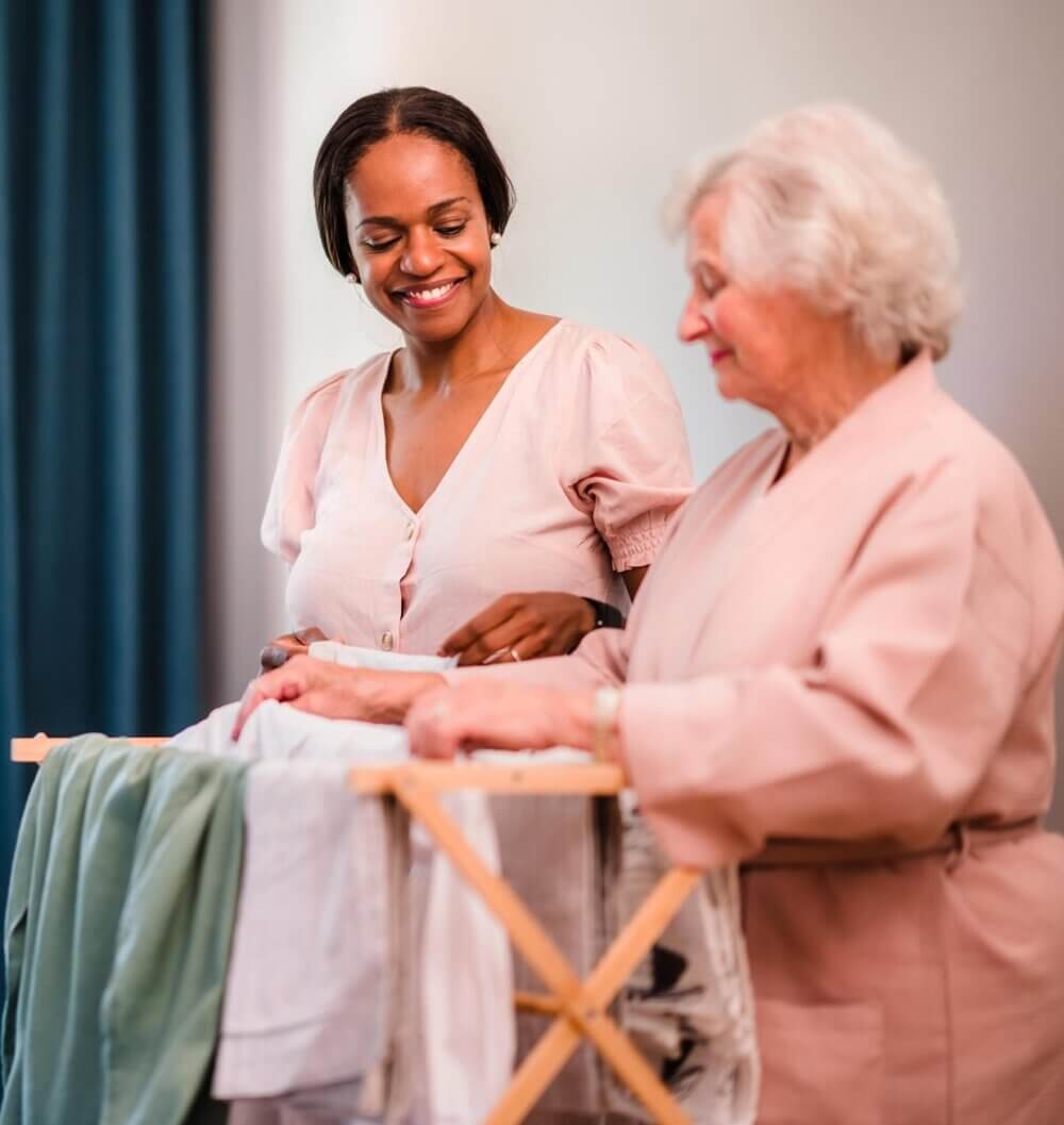 A younger woman helps an elderly woman fold laundry, both smiling and engaged in the activity together. - Home Instead