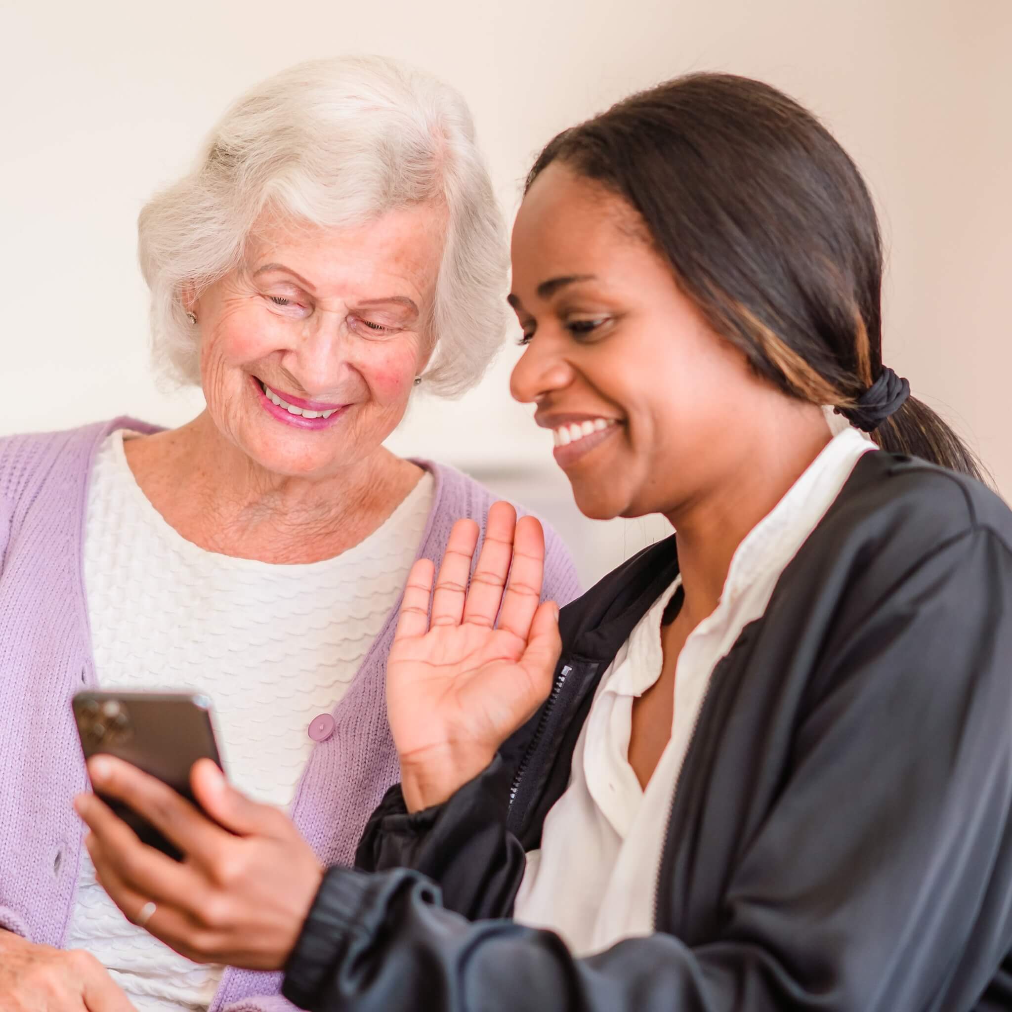 Two women smiling while looking at a smartphone, one elderly with white hair and the other younger with dark hair. - Home Instead
