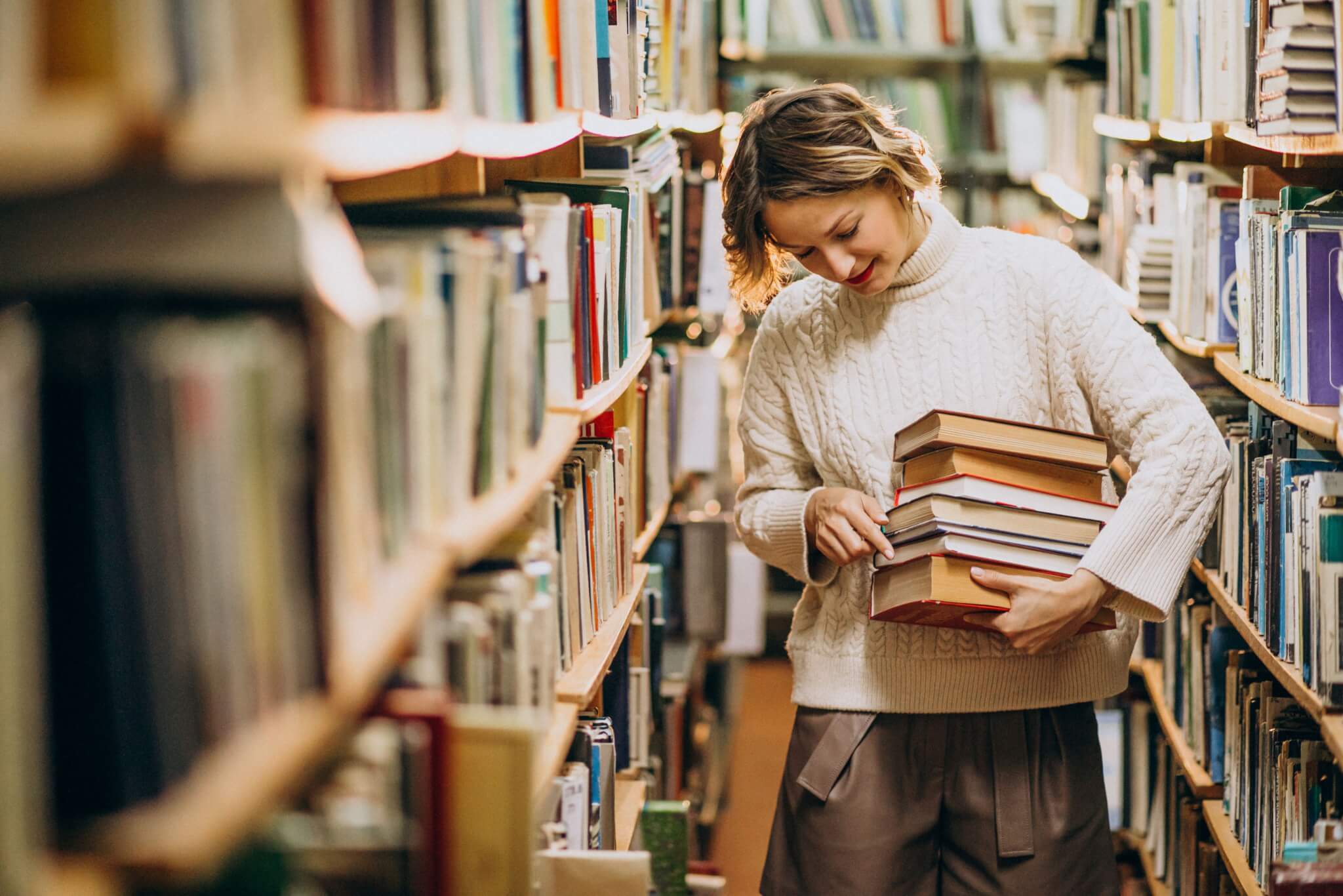 Person in a white sweater holding a stack of books in a library aisle filled with bookshelves. - Home Instead