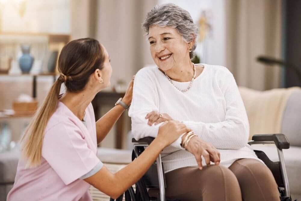 A smiling elderly woman in a wheelchair is talking to a Care Professional in pink scrubs who is holding her arm. - Home Instead Bournemouth & Christchurch