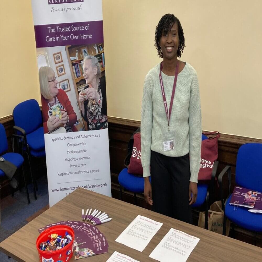 Woman standing at an information booth about elderly care services with pamphlets and promotional items on the table. - Home Instead