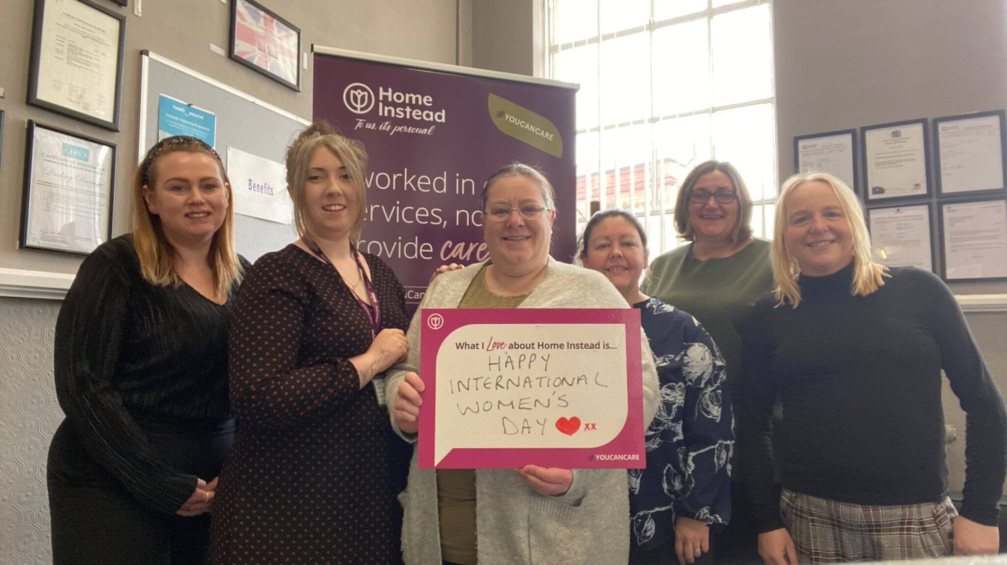 A group of six women smiling and holding a sign that reads "Happy International Women's Day" in an office setting. - Home Instead