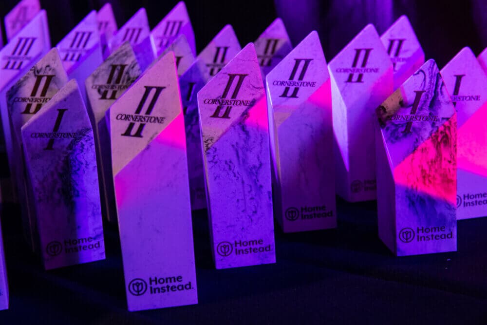 Several Cornerstone II awards with the Home Instead logo on a table, illuminated by purple light. - Home Instead