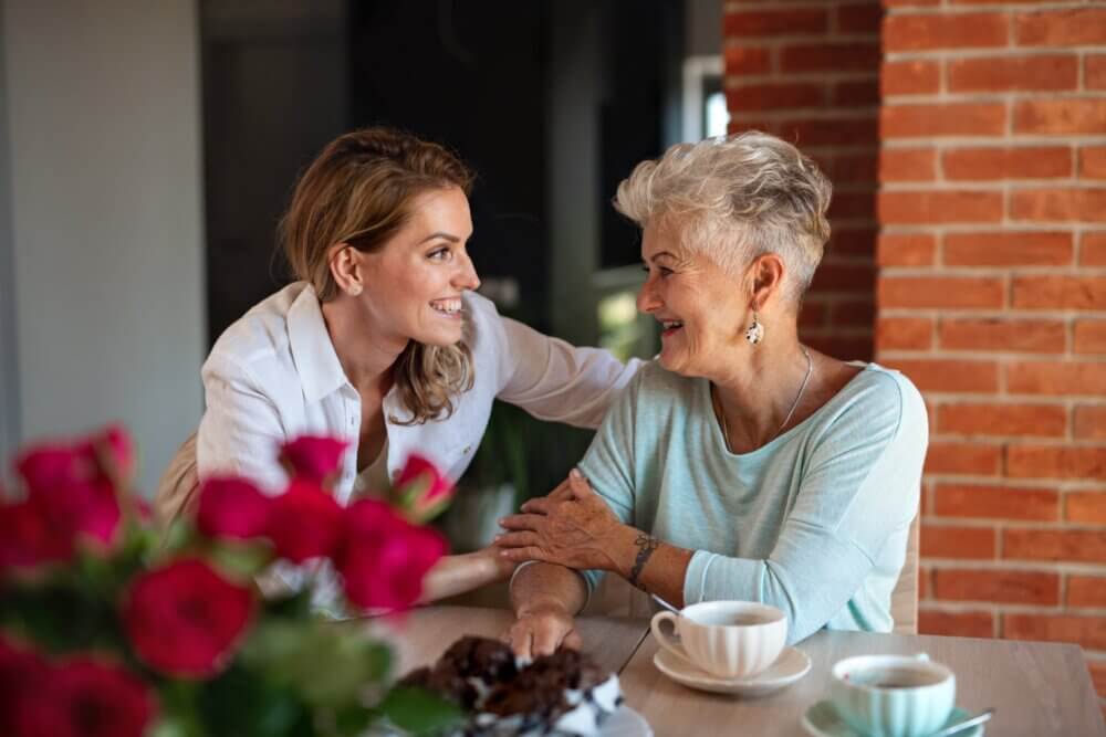 Two women, one younger and one older, enjoying coffee and conversation at a wooden table with a vase of red flowers. - Home Instead Southampton