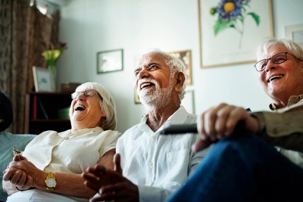 Elderly group with white hair laughing joyfully on a couch in a cosy room, with artwork on the wall behind them. - Home Instead Poole