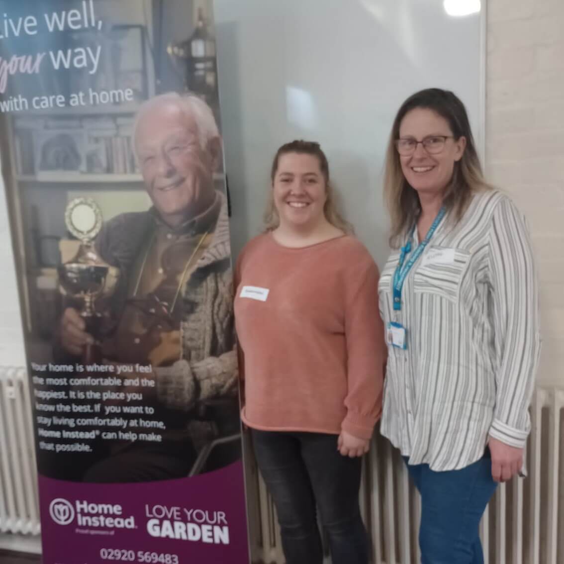 Two smiling women stand beside a care home banner offering home care services. - Home Instead