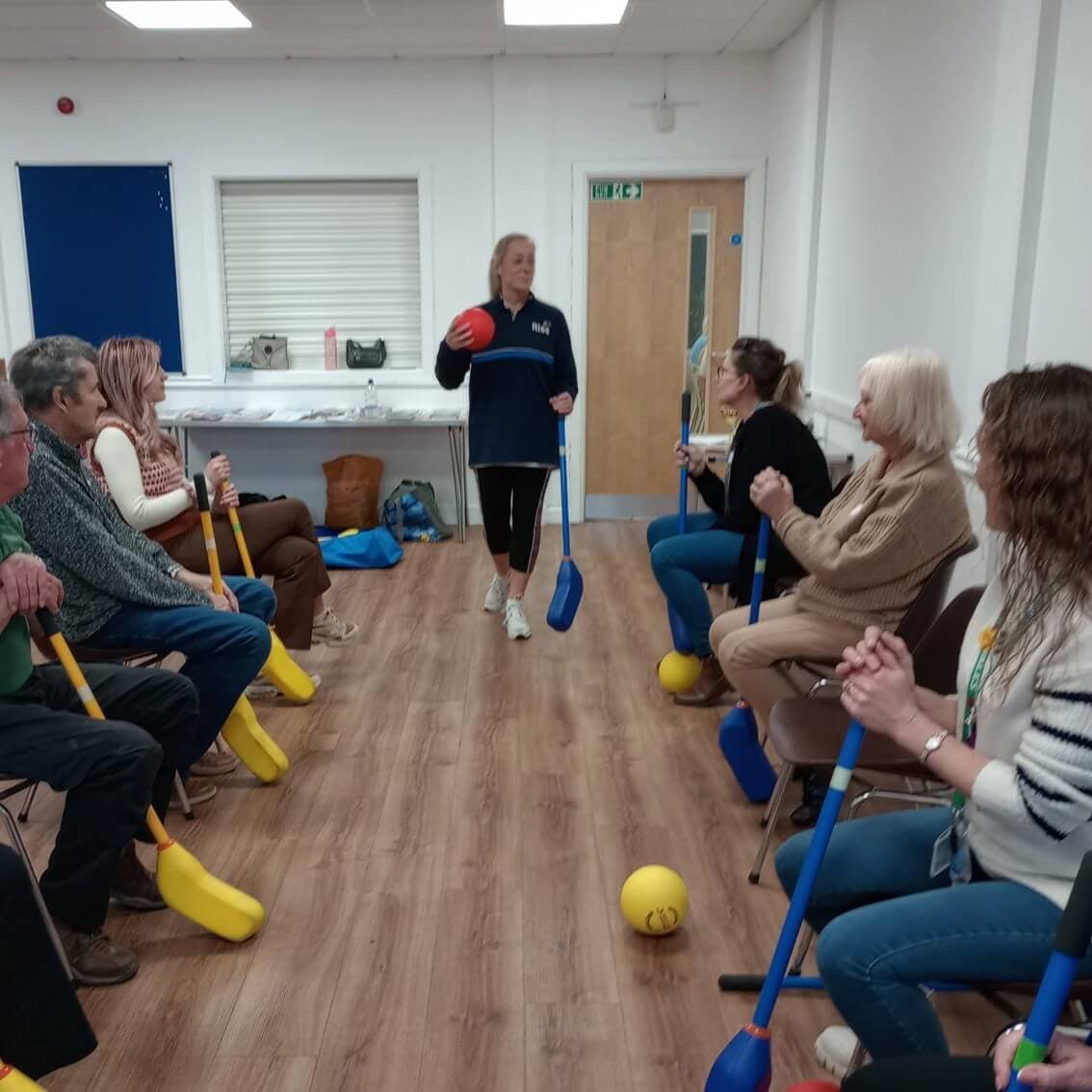 A woman instructs an exercise class with seated participants holding colorful sticks and balls in a room. - Home Instead
