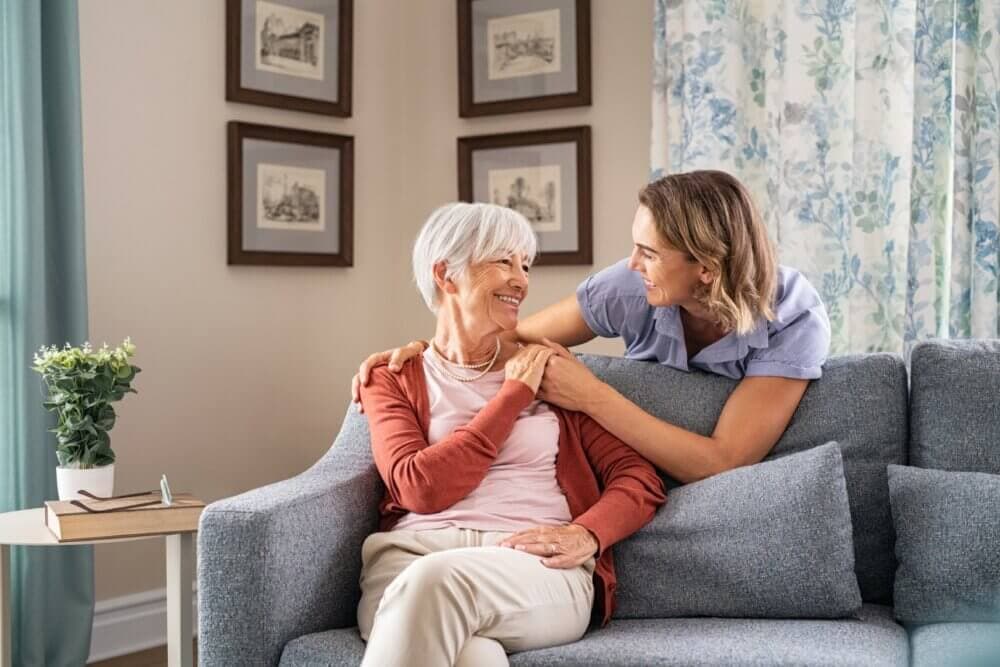 Elderly woman sitting on a couch smiling at a younger woman leaning over with hands on her shoulders. - Home Instead Southampton