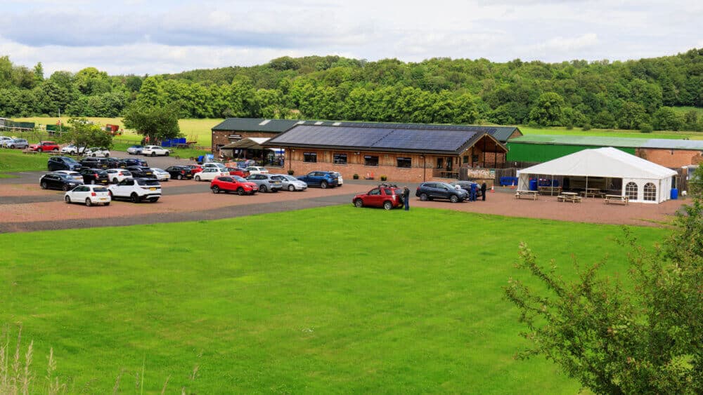 A rural parking lot filled with cars next to a large wooden building with solar panels and a white tented area. - Home Instead