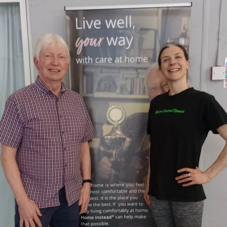 Two people standing in front of a poster with "Live well, your way" text; one is an older man, and the other is a younger woman. - Home Instead