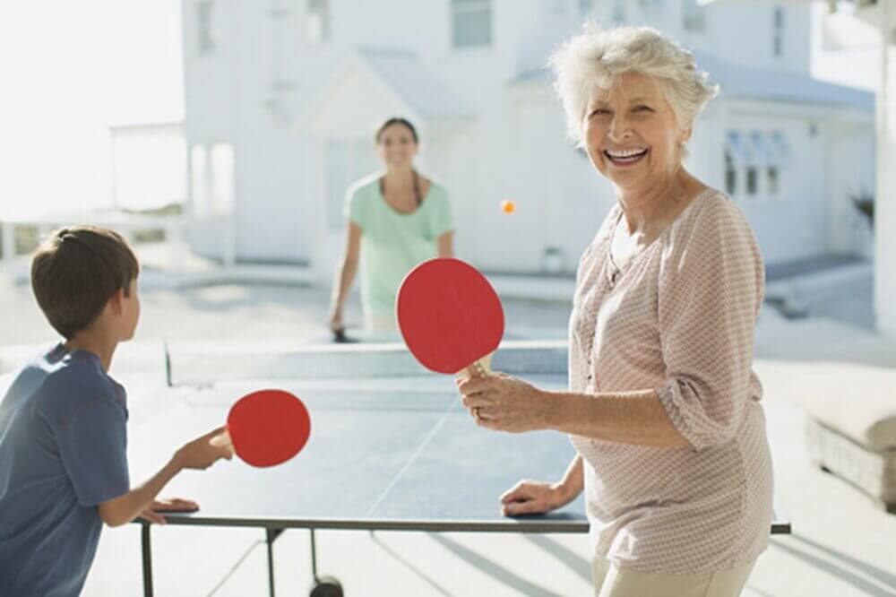 An elderly woman smiles while playing table tennis with a young boy and another woman outside on a sunny day. - Home Instead