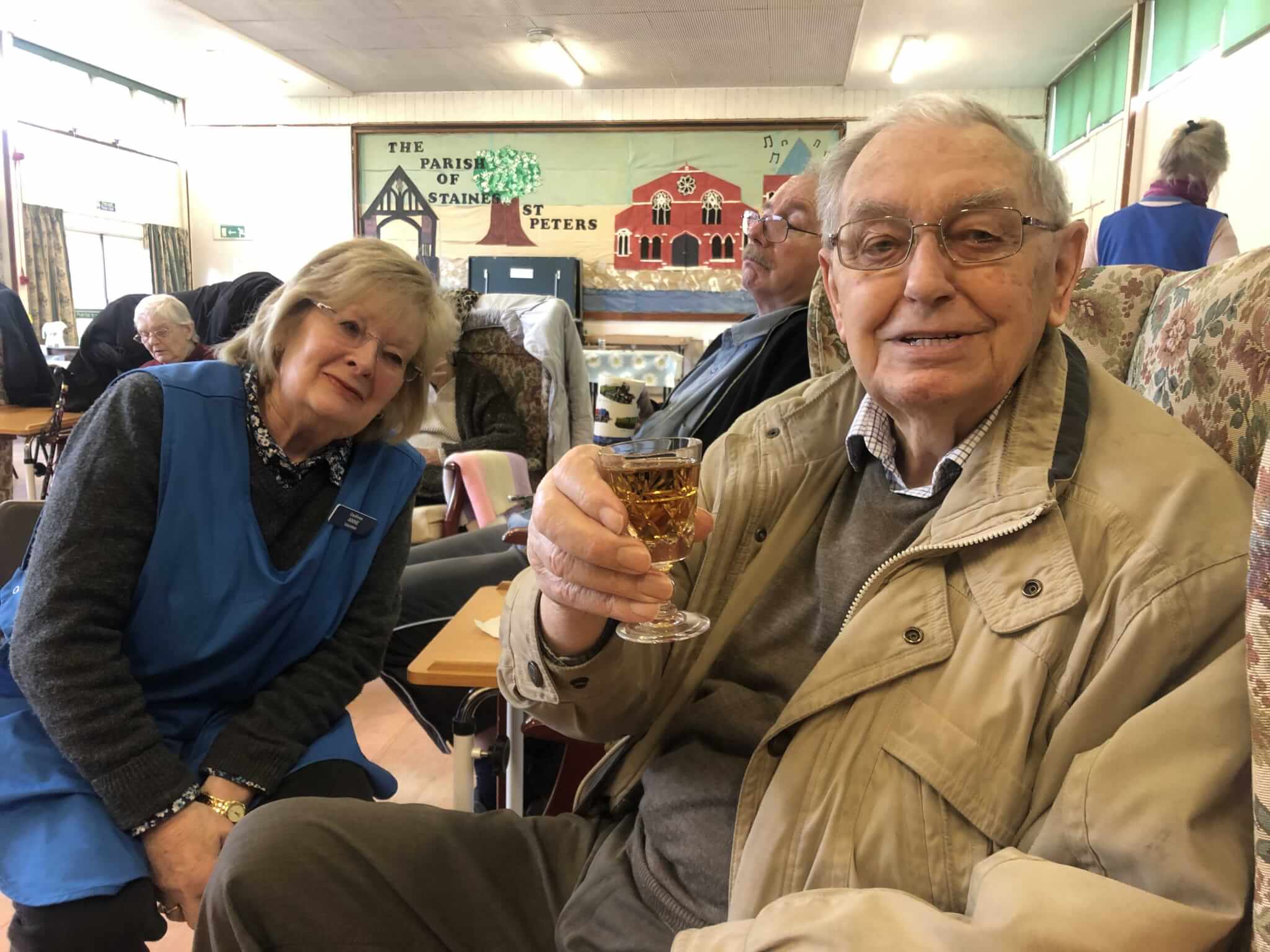 An elderly man with glasses holds a drink and smiles, while seated next to a woman in a blue vest in a communal setting. - Home Instead