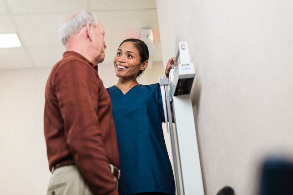 A healthcare professional in blue scrubs smiles while measuring a senior man's height in a medical office. - Home Instead