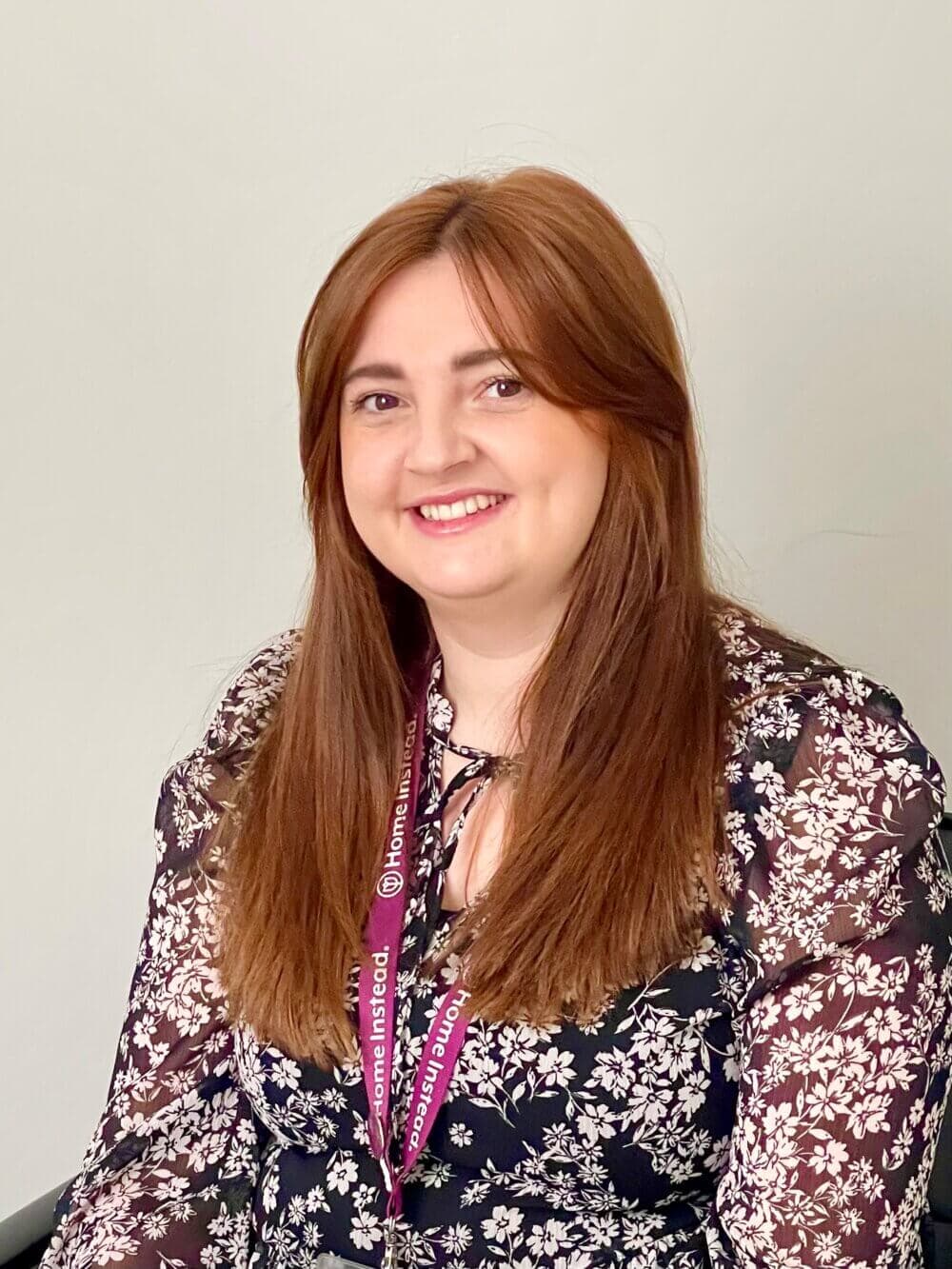 Woman with long brown hair wearing a floral blouse and lanyard, smiling against a plain background. - Home Instead