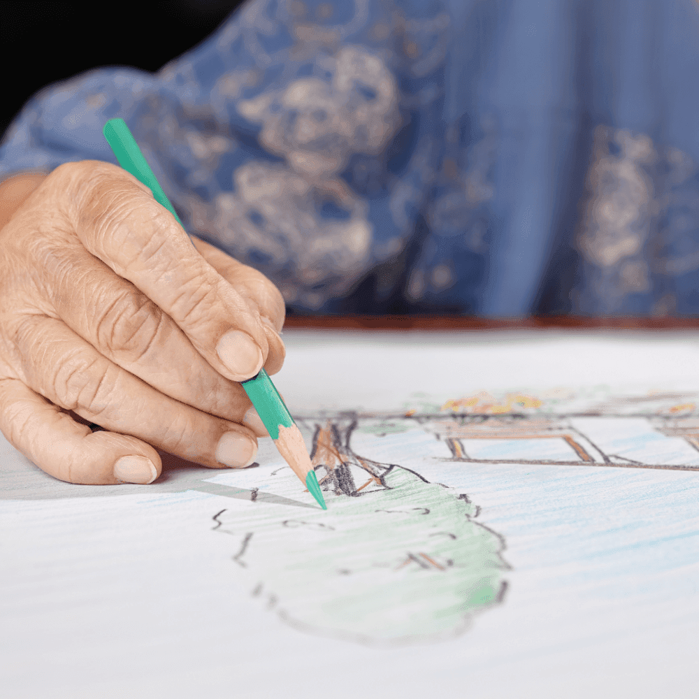 An elderly person with a green pencil is coloring a drawing, with part of a blue blouse visible in the background. - Home Instead