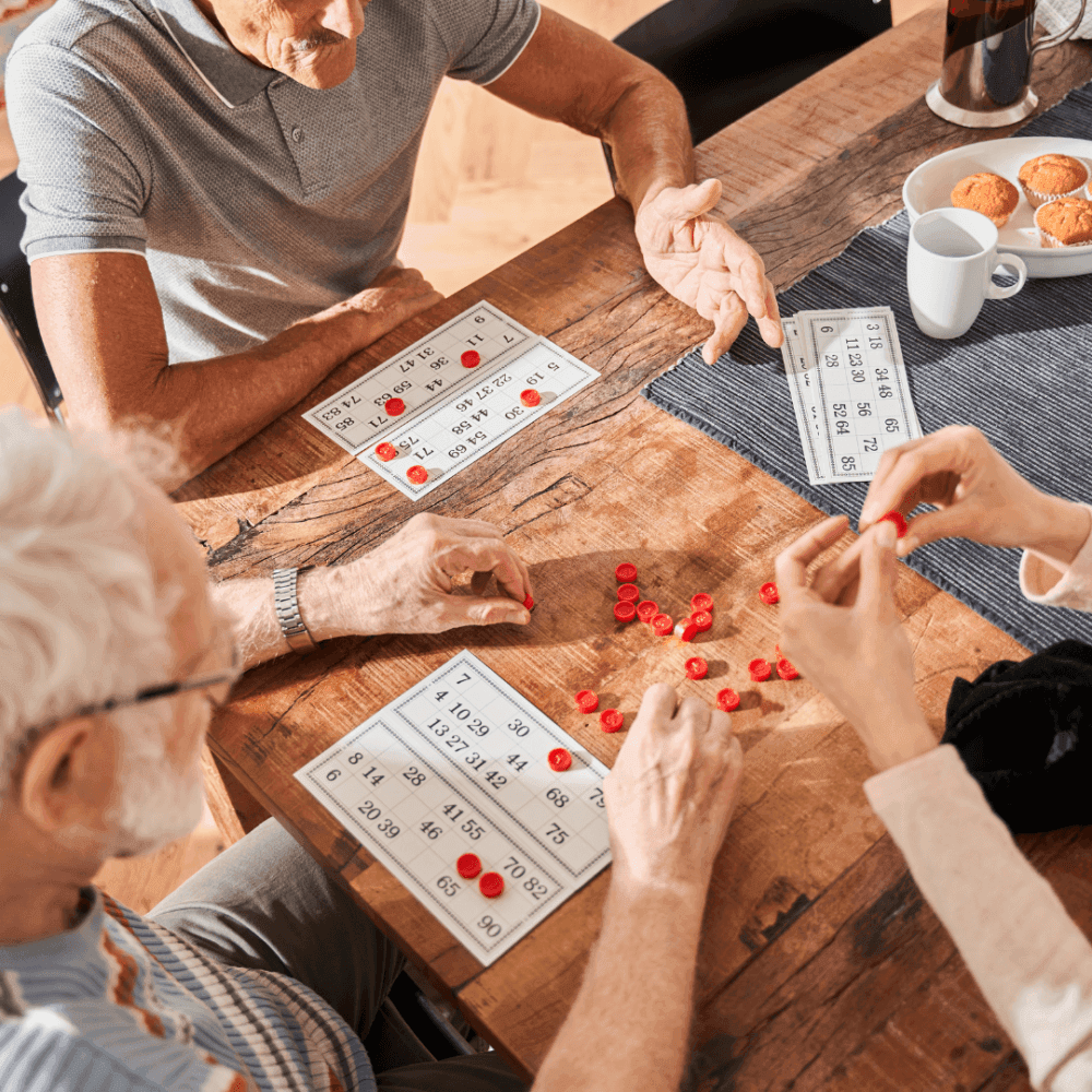 Three elderly people sitting around a table playing a game of bingo with red chips and bingo cards. - Home Instead