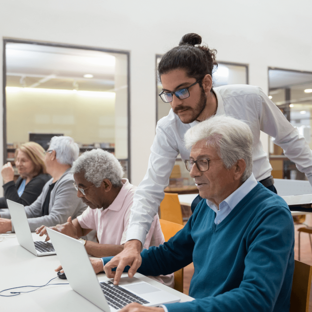 A young man assists a senior using a laptop in a classroom setting with multiple seniors working on computers. - Home Instead