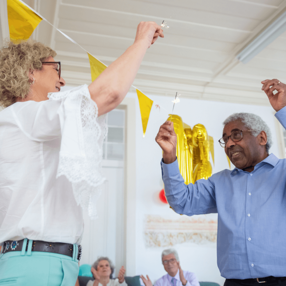 Two people holding sparklers while celebrating indoors, with yellow party decorations in the background. - Home Instead