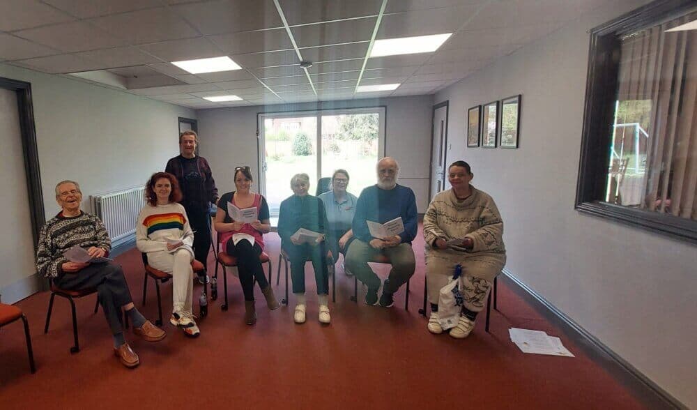 A group of eight people are sitting in a room, holding papers and smiling at the camera. - Home Instead