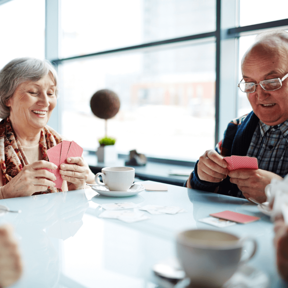 Two elderly people sitting at a table, smiling and playing cards. Cups of coffee are on the table in a bright room. - Home Instead