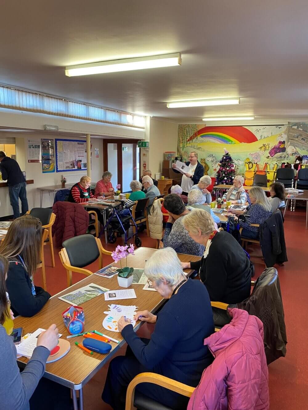 A group of elderly individuals engage in various activities in a community center decorated with colorful artwork and a Christmas tree. - Home Instead