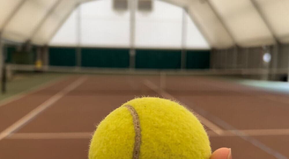 Close-up of a tennis ball being held up against the background of an indoor tennis court.