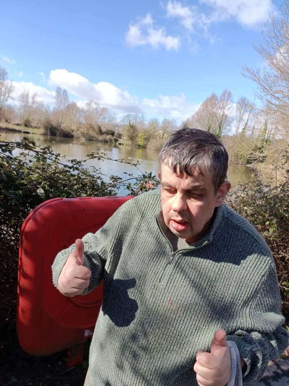 A man standing outdoors near a lake giving two thumbs up, with clear skies and trees in the background. - Home Instead