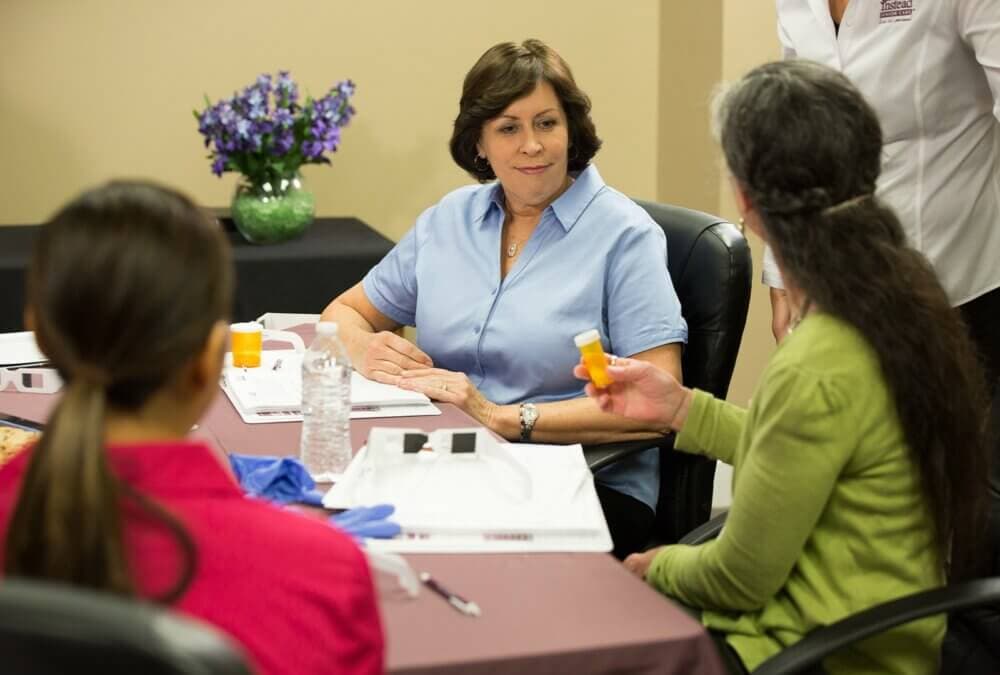 A woman in a blue shirt sits at a table, listening to a person holding a pill bottle, with others participating. - Home Instead