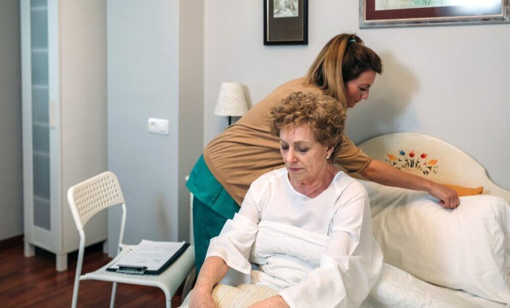 A Care Professional helps an elderly woman sit up in bed in a room with framed pictures on the wall and a chair by the bedside. - Home Instead Southampton