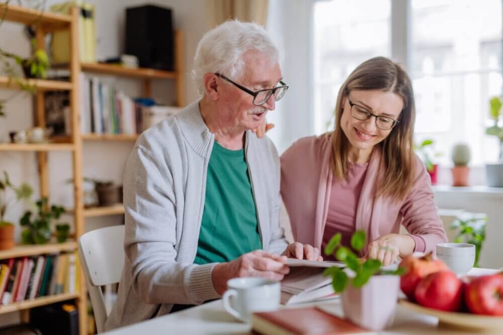 An elderly man and a younger woman read a book together at a table in a bright, cozy room with plants and bookshelves. - Home Instead Bournemouth & Christchurch